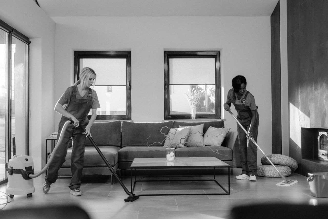 services-01 Two women in work clothes cleaning a modern living room using a vacuum and mop.
